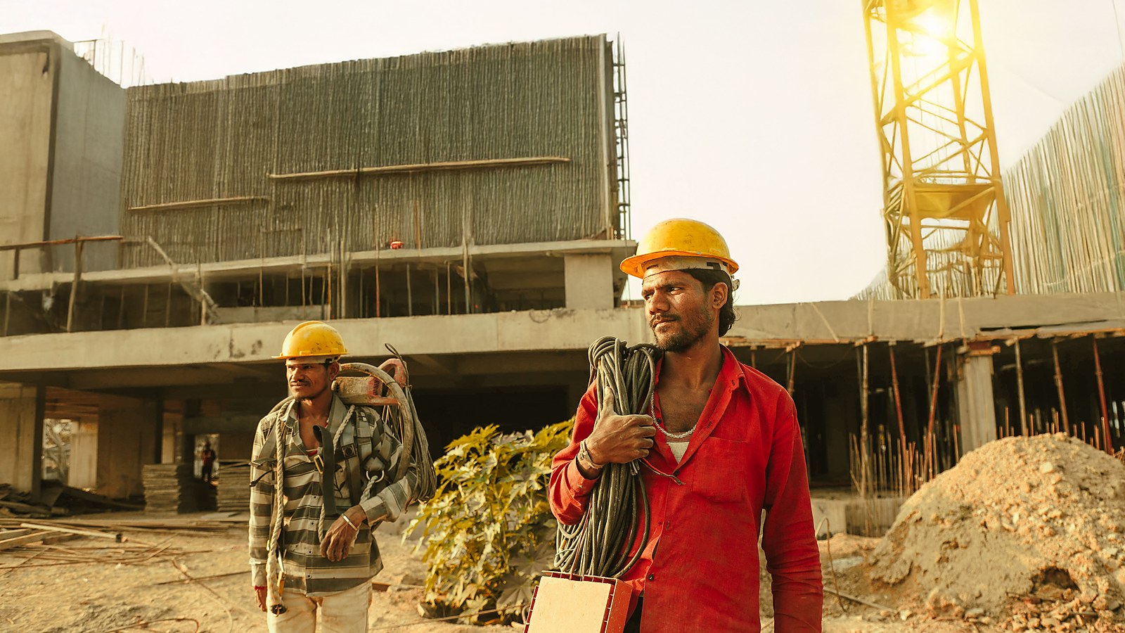 Operarios de capacete carregam cabos e material em canteiro de obras a ceu aberto, trabalhadores protegidos pelas normas de seguranca e saude no trabalho