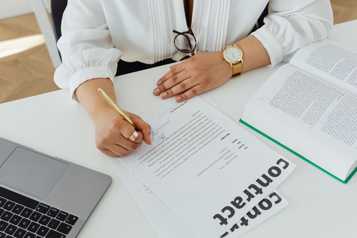 Profissional assina contrato de trabalho sobre a mesa, ao lado de um livro de direito