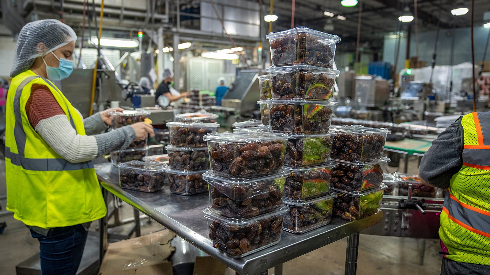 Trabalhadores de touca e máscara embalam alimentos em bancada de aço de uma linha de produção
