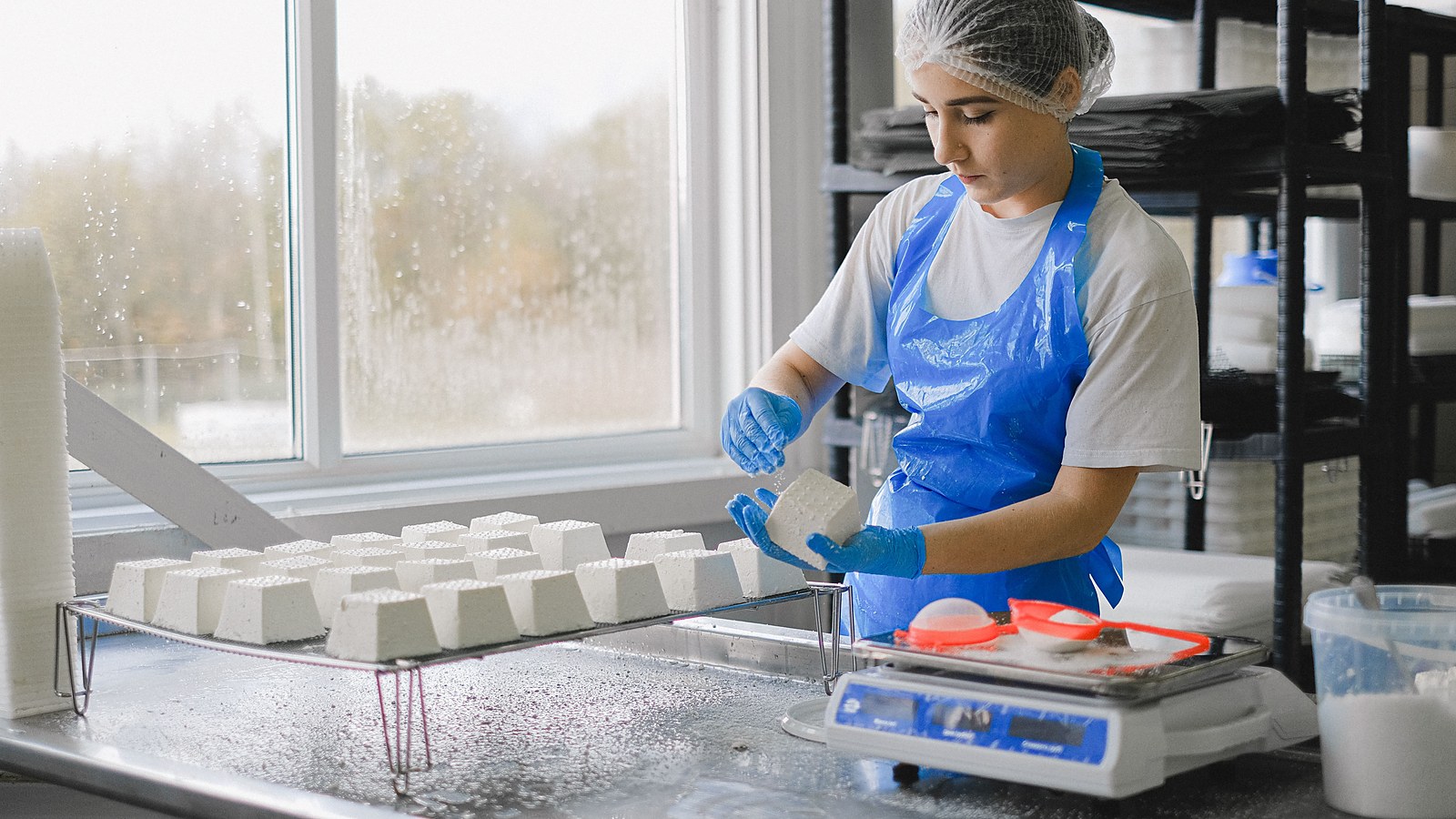 Trabalhadora de touca, avental e luvas manipula produto em bancada de aço inoxidável de indústria de alimentos