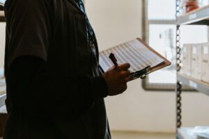 Profissional confere um formulário preso a uma prancheta em almoxarifado, com pilhas de documentos ao fundo